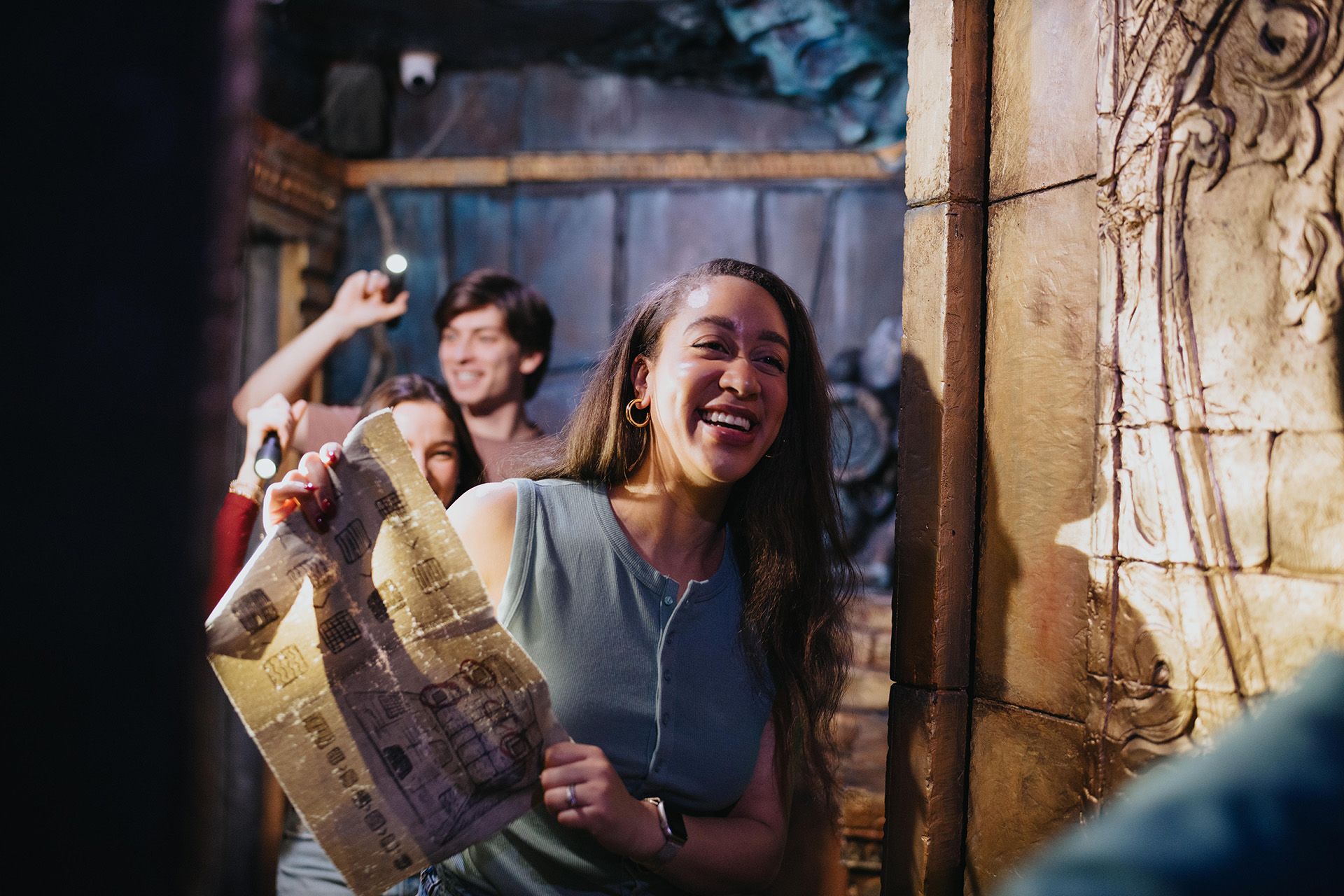 A smiling woman holds a map while standing in a dimly lit room with stone walls, surrounded by friends.