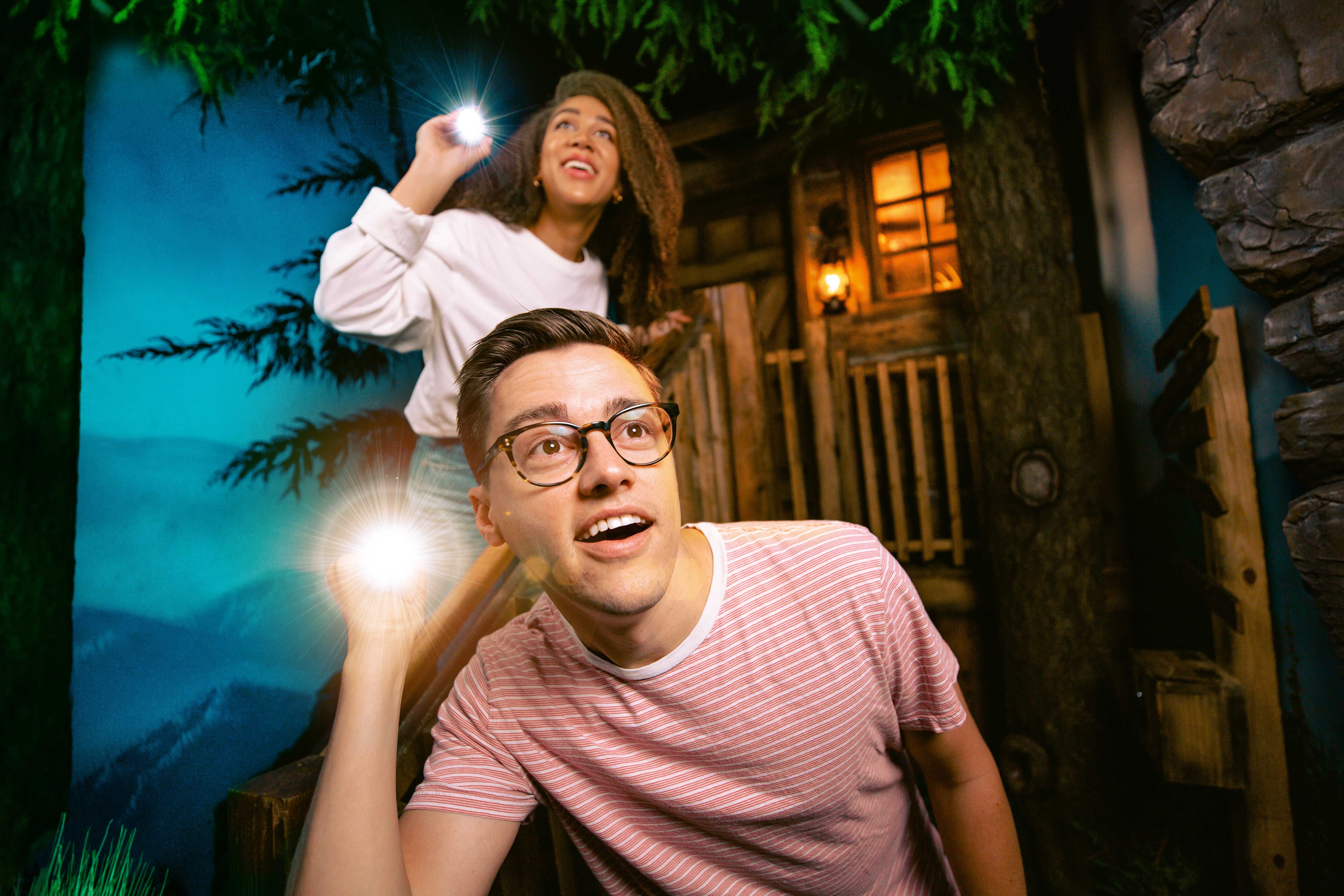 Two people explore a set resembling a forest cabin, holding flashlights and looking upwards with expressions of curiosity.