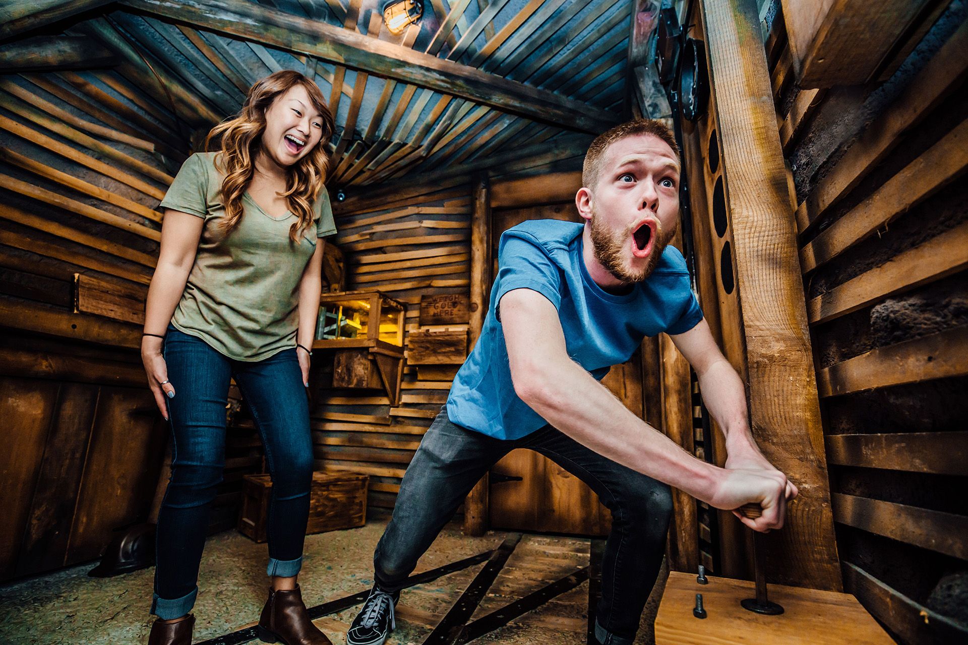 A man making an exaggerated surprised face while pulling a lever in a wooden room, with a laughing woman nearby.