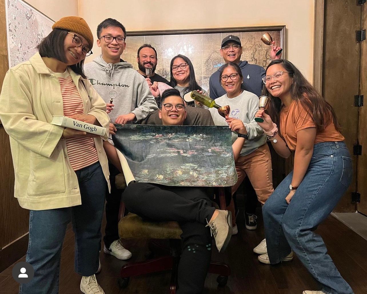 Group of eight people posing together, holding drinks and a painting. Smiling and having a fun time in a cozy indoor setting.