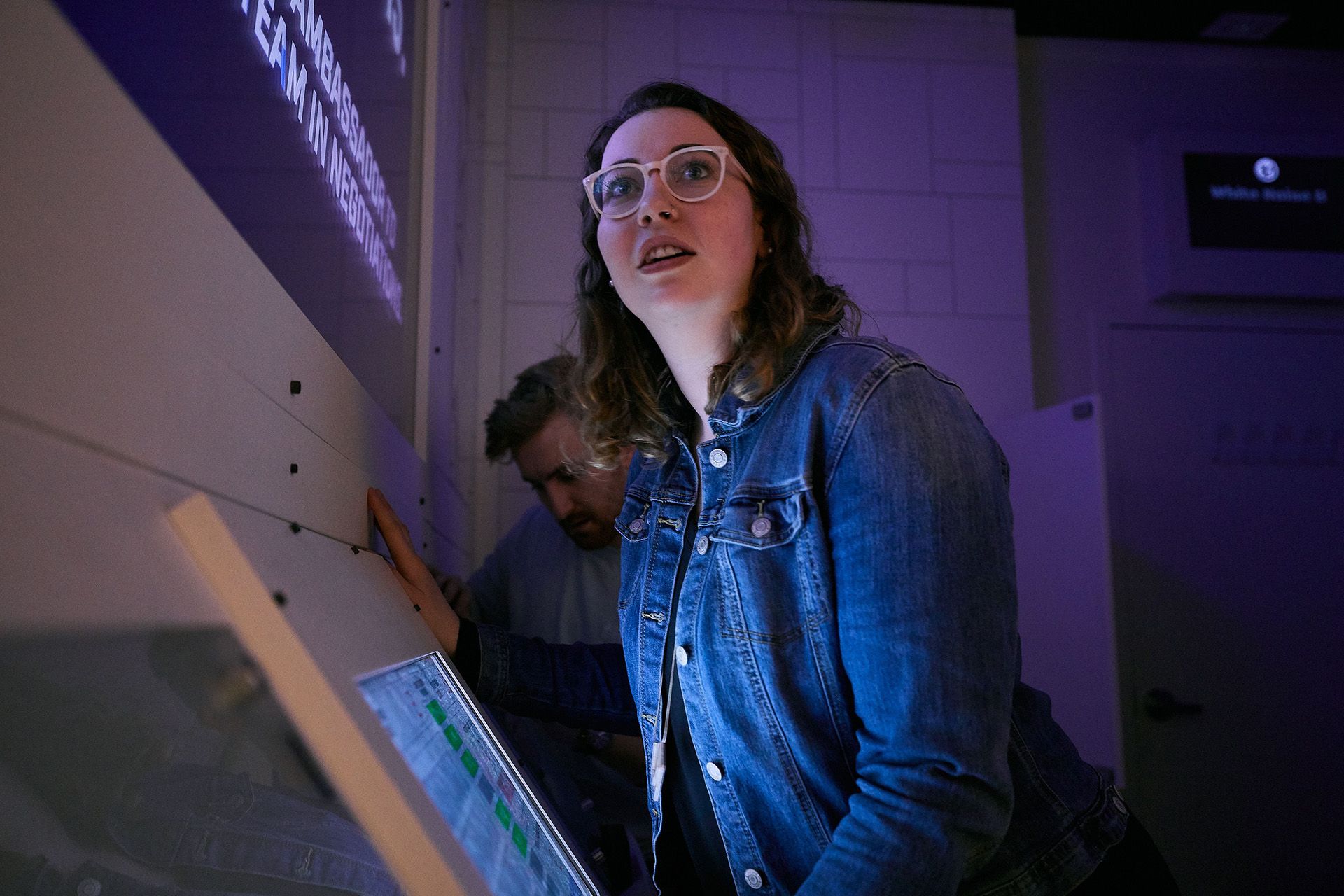 A woman in a denim jacket looks surprised while interacting with a touchscreen display, with a man working nearby in a dimly lit room.