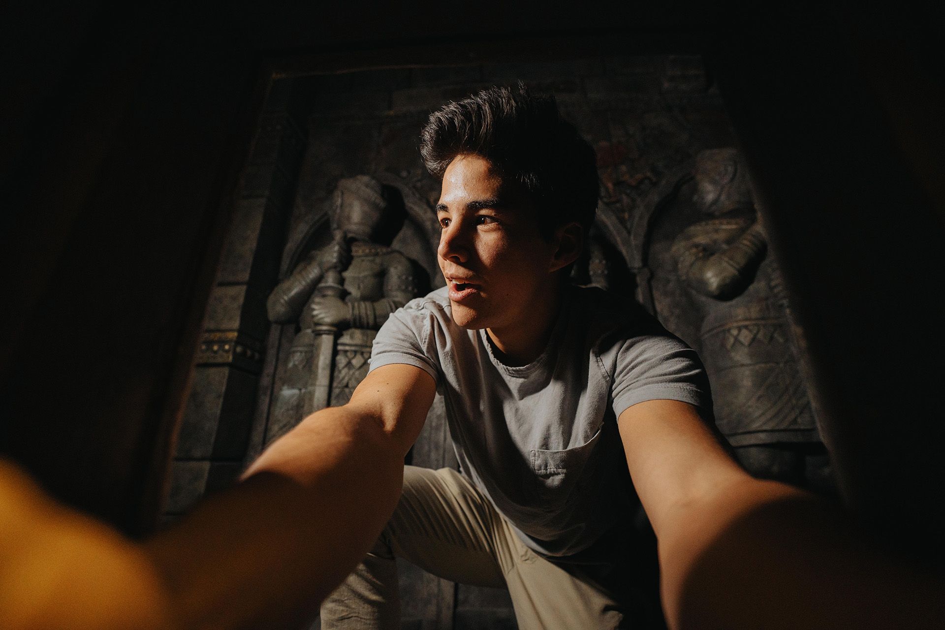 A young person in a gray shirt is taking a selfie in a dimly lit space with ancient stone statues in the background.
