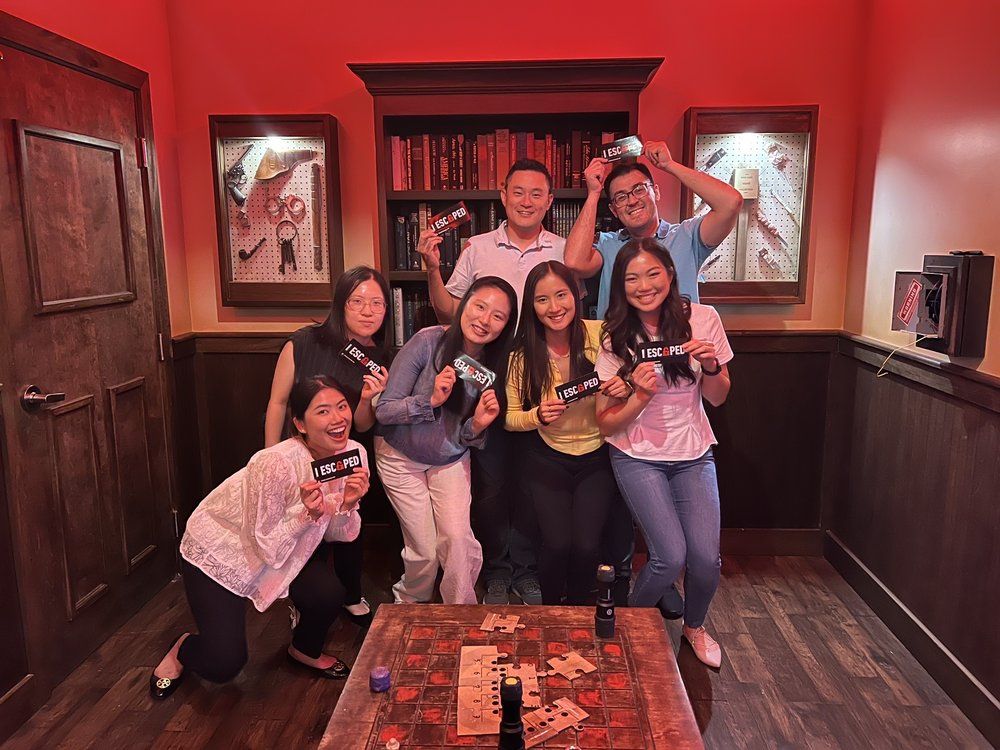 A group of seven people pose together, holding signs that say "I ESCAPED," in a room with red lighting and wooden decor.