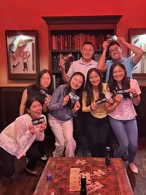 A group of seven people pose together, holding signs that say "I ESCAPED," in a room with red lighting and wooden decor.